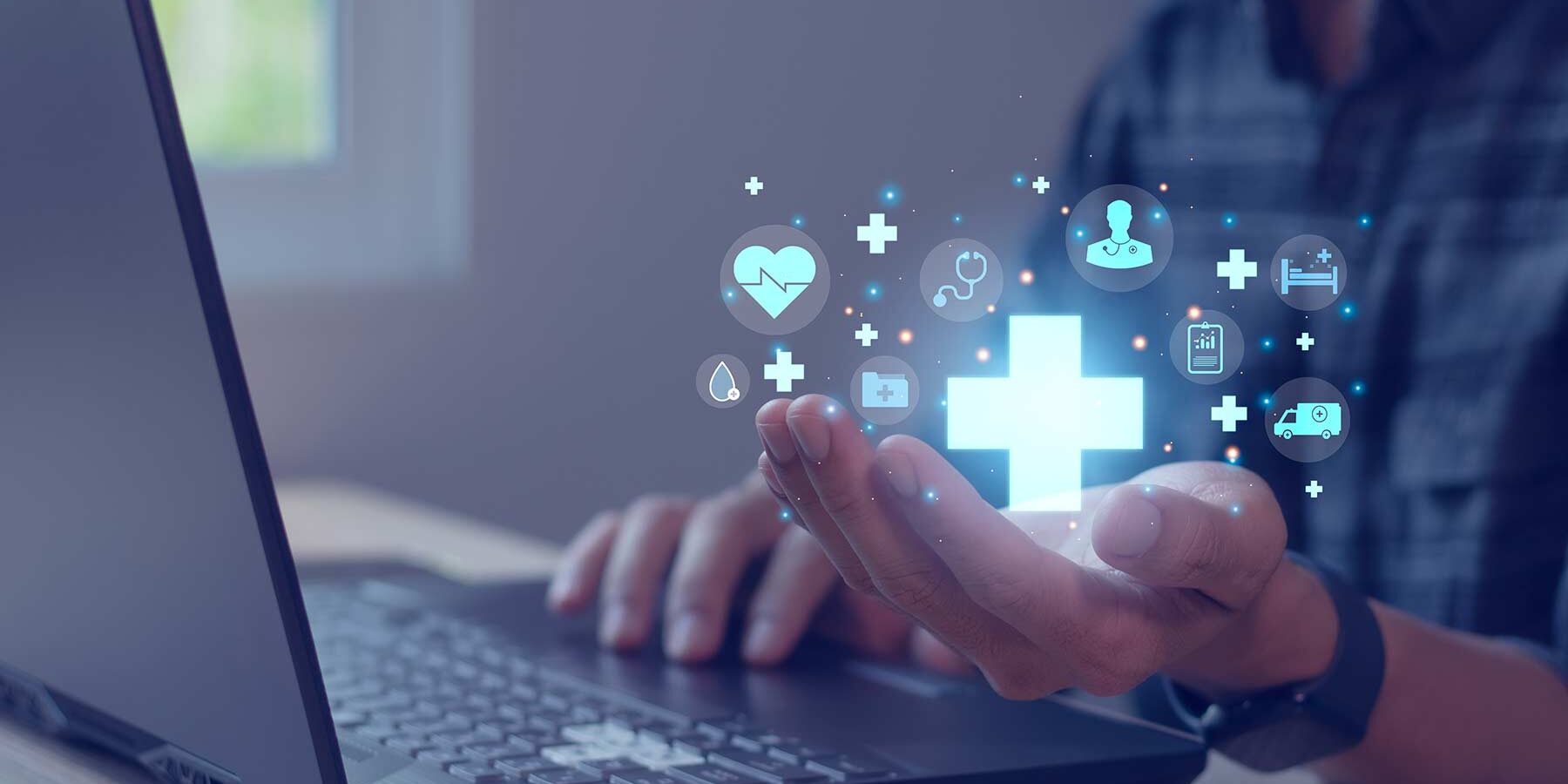Persönliche Hand über einer Laptop-Tastatur mit schwebenden blauen Symbolen, die Gesundheit und medizinische Dienstleistungen darstellen, wie Herz, Stethoskop und Krankenwagen, um ein zentrales Kreuz. Diffuses Tageslicht von links.