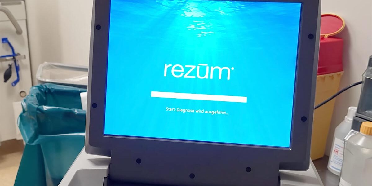 Ein Bildschirm zeigt das Wort "rezüm" auf blauem Hintergrund mit Wellenmuster. Darunter steht "Start-Diagnose wird ausgeführt...". Der Bildschirm gehört zu einem medizinischen Gerät in einem Raum mit Behältern im Hintergrund.